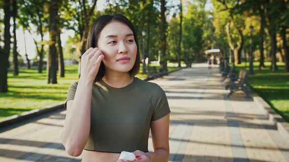 Outdoor Portrait of Young Asian Lady Putting in Wireless Earphones in Park and Smiling Tracking Shot