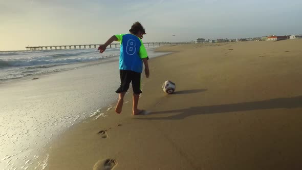 A boy kicks a soccer ball on the beach at sunset with the ocean and pier.