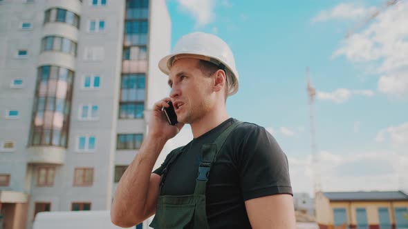 A Builder Wearing Helmet Is Talking By Phone While Inspecting Building Site.