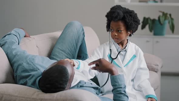 Cute Little Girl in Medical Gown Plays with Dad Pretending to Be Doctor Man Lying on Sofa Funny