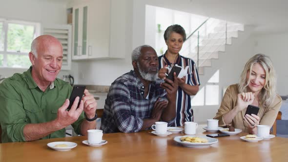 Two diverse senior couples sitting by a table drinking tea using smartphones at home