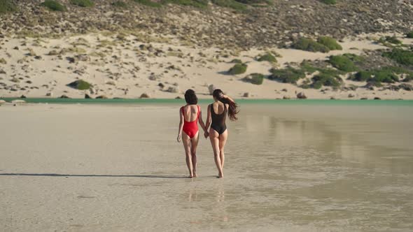 Pretty Female Friends Walk Holding Hands By Sea on Sandy Tropical Beach in Swimsuits