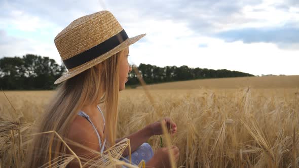 Thoughtful Little Girl in Straw Hat Sitting in the Wheat Field and Touching Spikelet. Beautiful