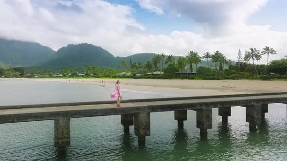 Aerial Lady in Dress Walking By Beach Pier with Cinematic Mountain Landscape