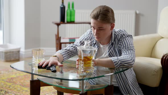 Portrait of Desperate Young Man Moving Apart Glass and Decanter with Whiskey Taking Gun in Hand and