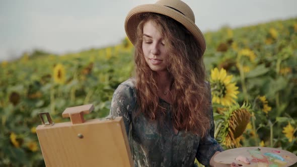 Longhaired Lady Paints with Watercolours Among Sunflowers in Windy Day