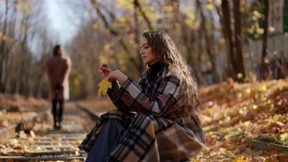 a Woman with Curly Hair and a Plaid Coat is Sitting on the Rails with a Yellow Leaf in Hands on a