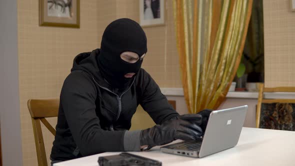 A Robber Is Trying To Hack Into a Laptop. A Masked Thug Is Sitting in a House and Cannot Hack