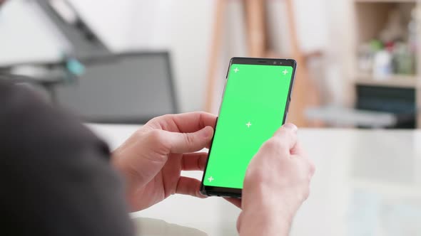 Man Sits at a Desk Holds His Smartphone with Green Screen on