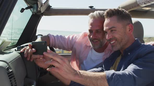 Happy caucasian gay male couple taking selfies sitting in car smiling on sunny day at the beach