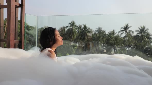 An asian woman taking a bubble bath in a jacuzzi on a balcony and taking a deep breath relaxing with