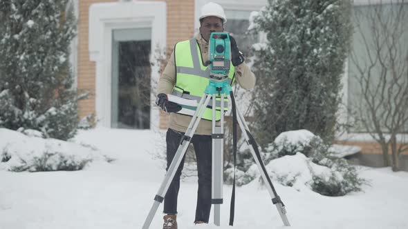 Wide Shot Portrait of Professional Concentrated African American Man in Hard Hat Using Theodolite