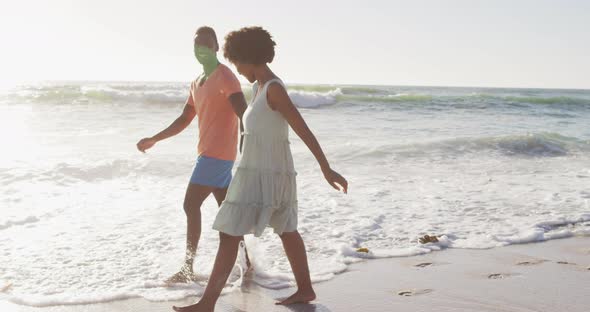 Smiling african american couple holding hands and walking on sunny beach