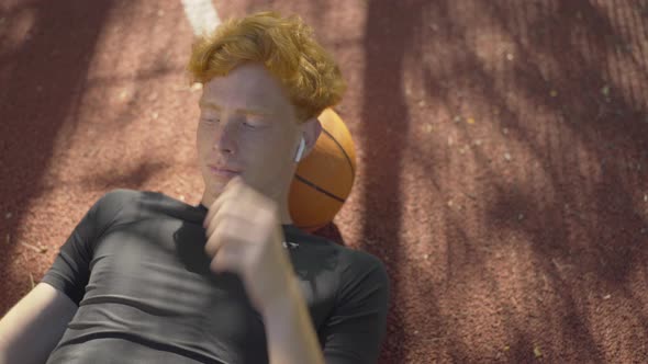 Top View of Young Redhead Caucasian Man Lying on Basketball Ball on Outdoor Court and Listening To