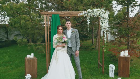 Camera Approaches Bride and Groom. Girl with Bouquet Stands Near Beloved Husband