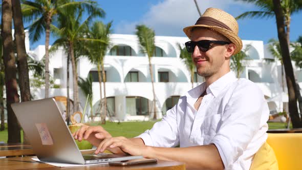 Digital Nomad Man In Hat Working Remotely On Laptop On Beach