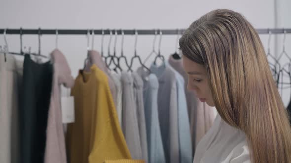 Side View Smiling Slim Young Woman Examining Clothes in Shop Thinking