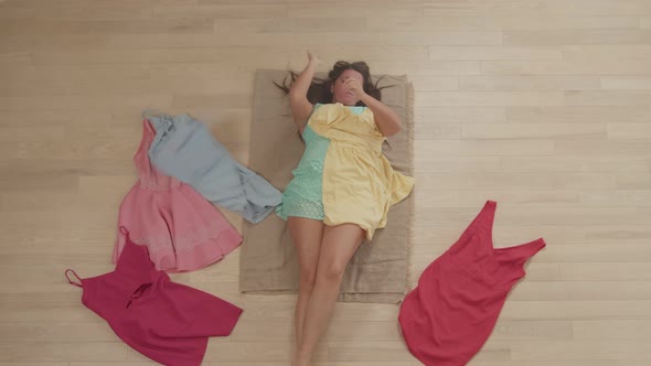 Top Angle View of Plump Woman Choosing Dresses Lying on Floor Indoors