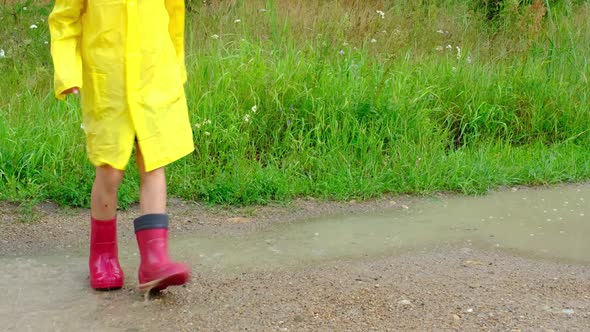 A girl in red rubber boots and a yellow raincoat runs through puddles after a rain in the village. S