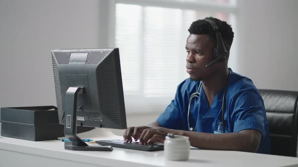 A Black Man Sits at a Computer in a Doctor's Uniform and Writes a Patient's Card While Taking Calls