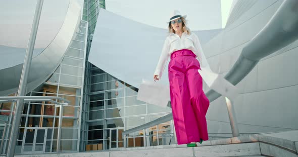 Happy Woman in Fashionable Clothes Dances with Shopping Bags at Modern Building