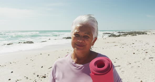 Portrait of hispanic senior woman standing on beach, holding yoga mat and smiling