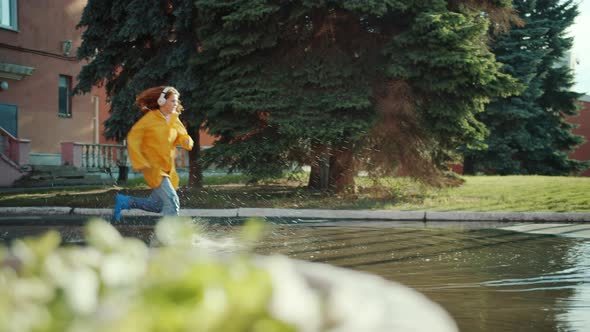 Girl in Raincoat and Gumboots Running in Puddles Wearing Headphones Having Fun