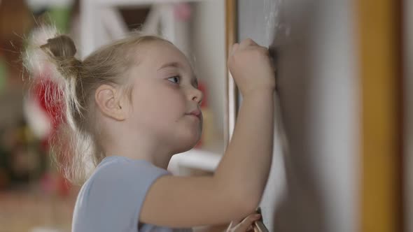 Side View of Genius Little Girl Writing with Chalk on Blackboard in Classroom
