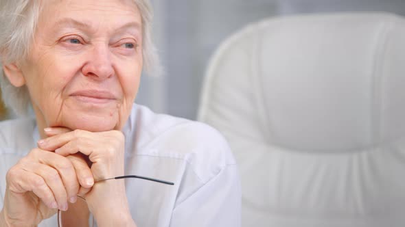 Aged woman with short grey hair looks out of window holding glasses in hand