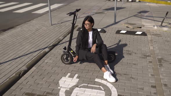 Brunette Business Woman in Suit Sitting on Escooter at Electric Car Charging Parking Lot