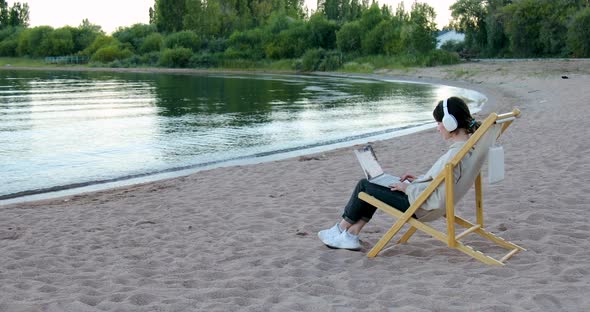 Freelance woman in headphones sitting sideways working on laptop by the lake