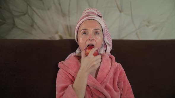 Woman Watching a Late Night Movie at TV, Eating an Apple. Bathrobe, Facial Mask