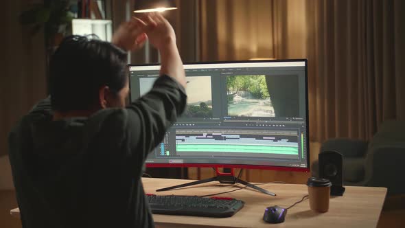 Asian Male Video Editor Stretching While Works With Footage And Sound On His Personal Computer