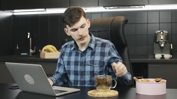 Caucasian man with mustache drinking tea during online work on laptop on kitchen. Work from home