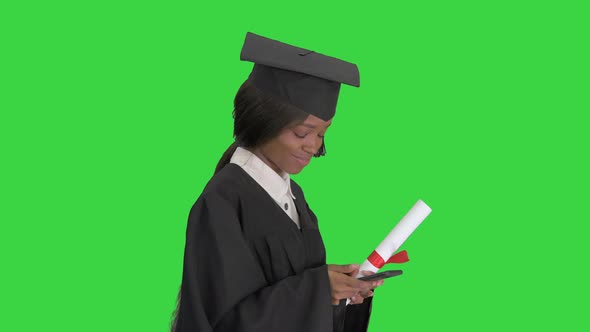 Smiling African American Female Graduate with Diploma Texting on Her Phone While Walking on a Green