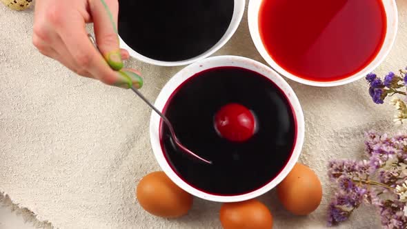 Caucasian Child Paints an Easter Egg with a Metal Spoon and Dips It Into a White Bowl with Red Dye