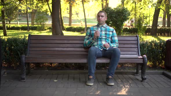 A Young Man Sits on a Bench in the Park and Drinks Cold Coffee Closeup. In the Background Nature