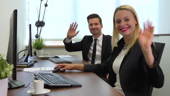 Two office workers, man and woman, work on computers and wave their hands at the camera, smiling