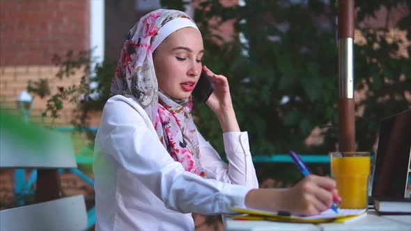 A Muslim Woman Makes an Important Call on Mobile Phone Makes Notes in a Notebook