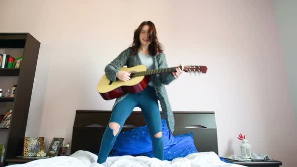 Young woman playing guitar jumping bed