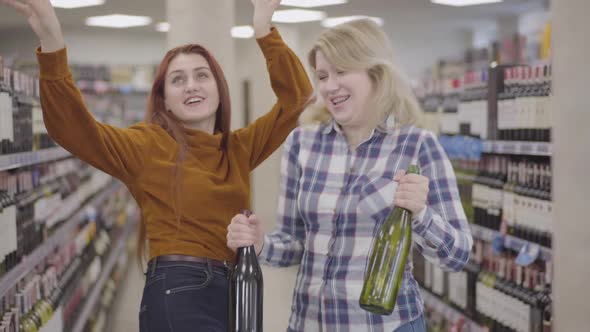 Happy Adult Caucasian Women Dancing in Wine Shop with Bottles of Beverage in Hands. Cheerful Blond