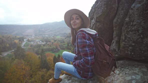 Girl Hiker in Checkered Jacket and Hat with Backpack Sits on Edge of Cliff and Enjoing Beautiful