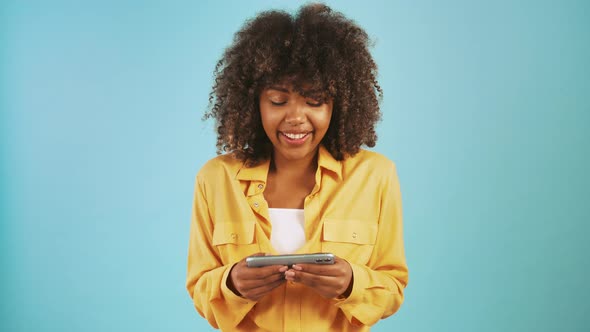 Afro American Woman Playing Video Game on Smartphone and Smiling While Posing Against Blue Studio