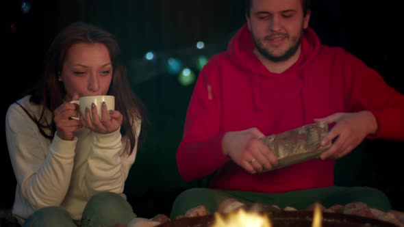 Couple of Travelers Bask By Fire in Campsite. A Man Throws Firewood in a Fire