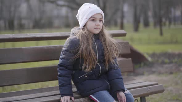 Brunette Schoolgirl Sitting on Bench in Park and Raising Leg. Portrait of Lonely Sportive Kid