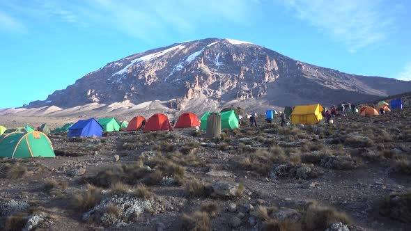 Wide shot of Mount Kilimanjaro Summit with Blue Sky. Multiple Tents and People in foreground.