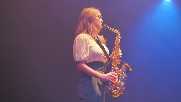 Young woman musician playing solo on saxophone under stage lights in haze