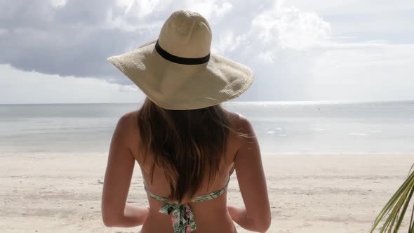 Camera following young woman with bikini and hat walking on beautiful beach towards clear water in u