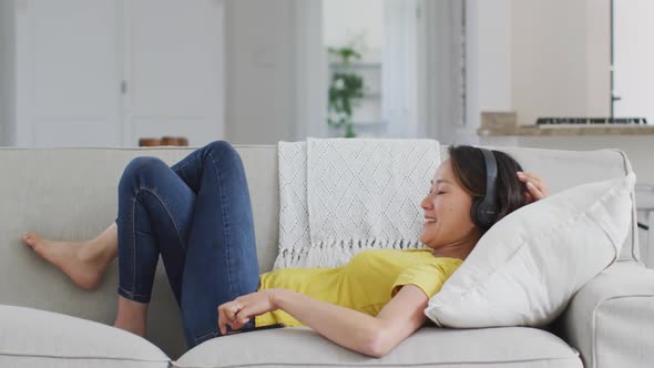 Happy asian woman wearing headphones lying on sofa, resting with smartphone at home