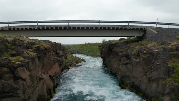 Aerial View Drone Flying Under Bridge Over Flowing River in Rocky Countryside in Iceland